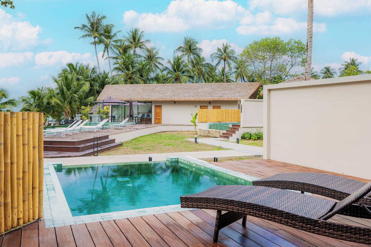 A pool area with lounge chairs, a thatched-roof building, and palm trees.