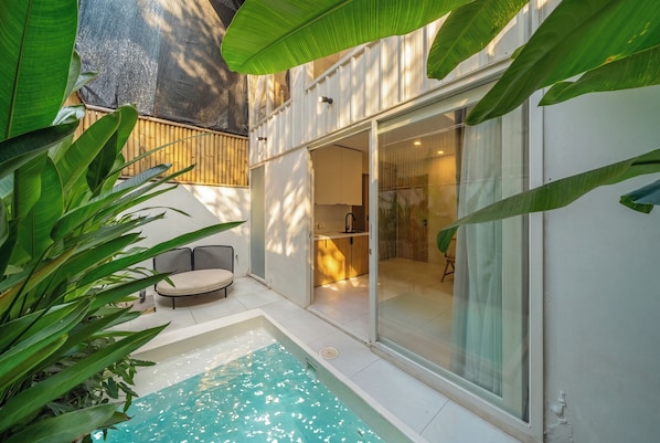 A modern outdoor pool area with a glass door leading to a room, surrounded by lush greenery.