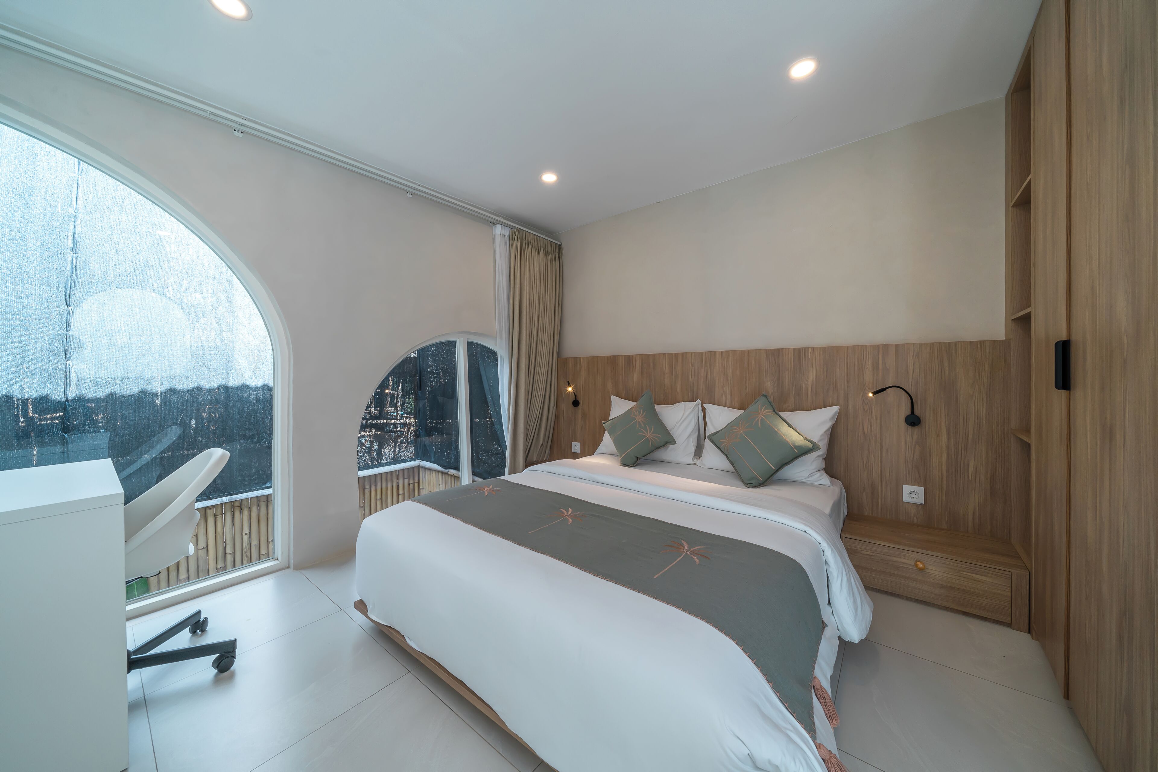 A modern bedroom with a large bed, a desk, and a view of a cityscape through a large window.