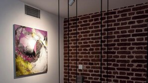 A room with a brick wall, a hanging abstract painting, and a visible vent. - The Nanee (Bhaktapur)