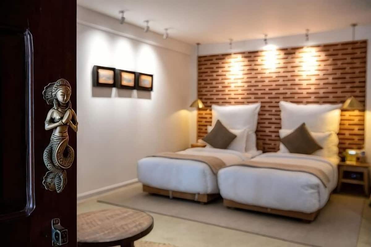 A bedroom with a brick-patterned headboard, two beds with white linens, a round side table, and wall-mounted picture frames.