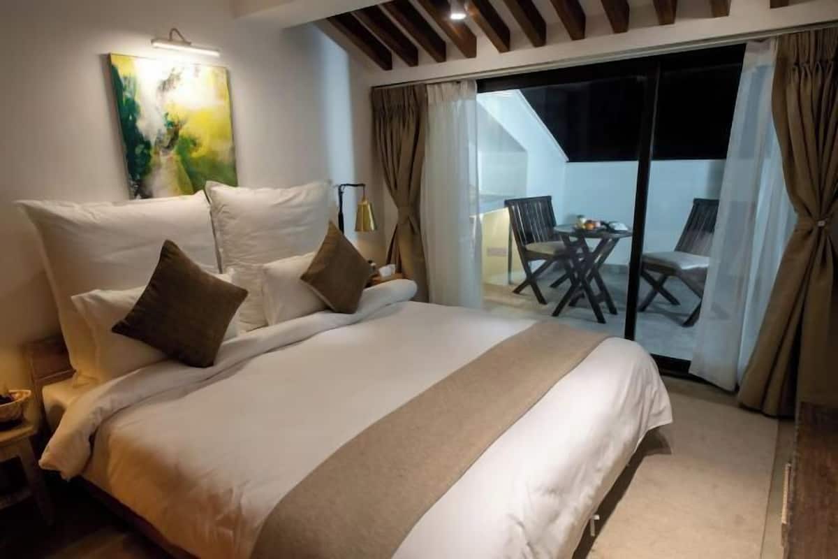 A large bed with white linens and a beige accent, a painting on the wall, and a balcony with a table and chairs.