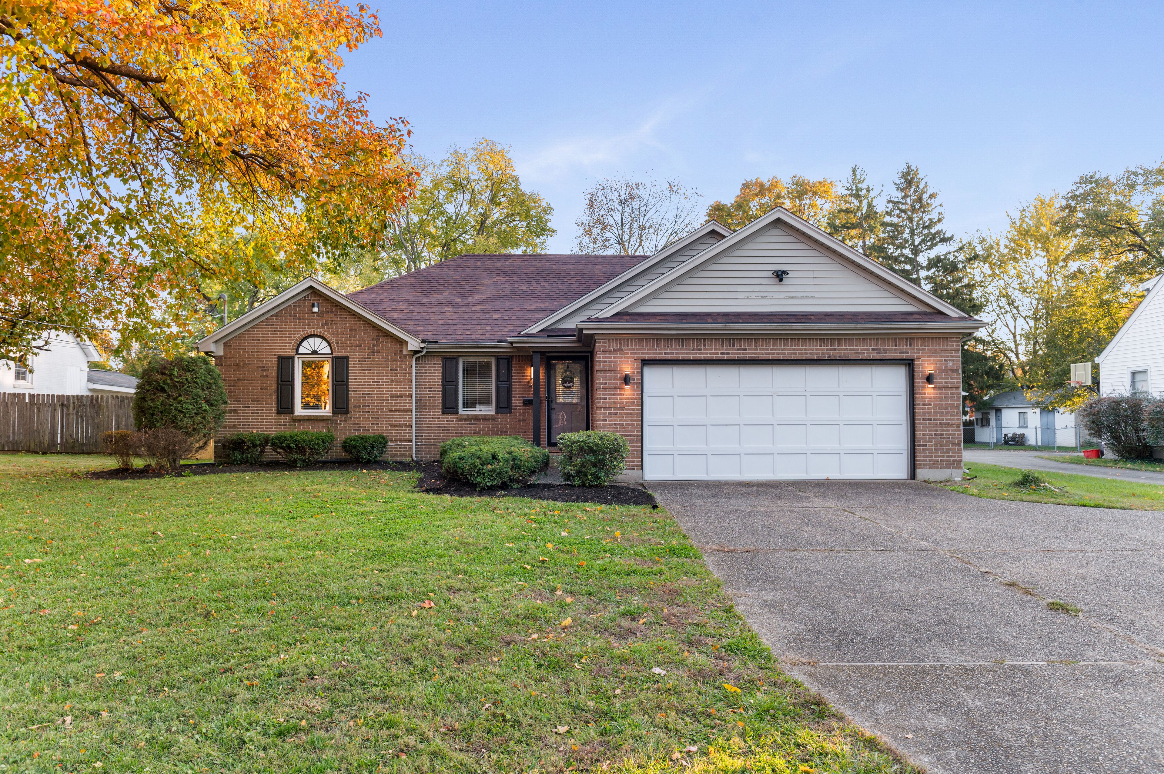 A single-story brick house with a garage, a front yard with grass and a small tree, and autumn-colored leaves on the trees.