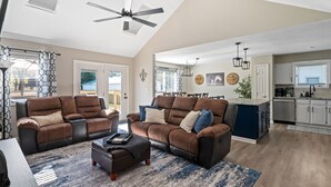 A spacious living room with a large brown sectional sofa, a coffee table, and a ceiling fan. There is a kitchen area with white cabinets and a dining area with a table and chairs.