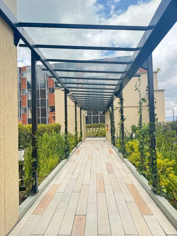 A covered walkway with a glass roof, bordered by plants and buildings.