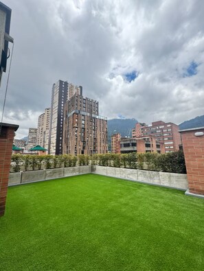 A rooftop with artificial green grass, surrounded by high-rise buildings and a cloudy sky.