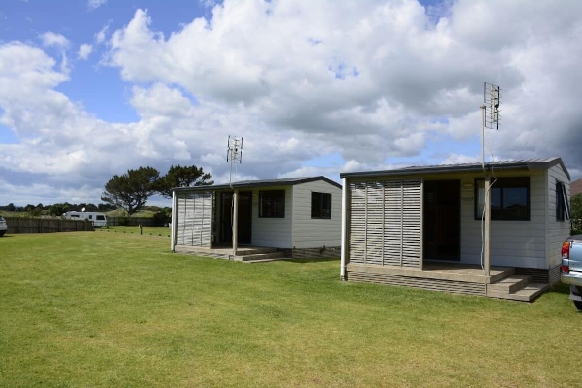 Two small, single-story mobile homes with external antennas, set in a grassy area with a clear sky and a few clouds.