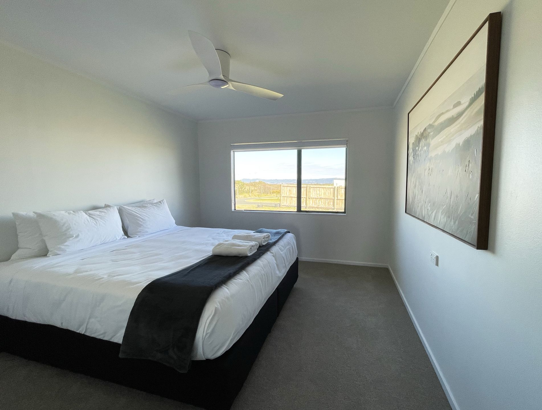 A bedroom with a bed, a ceiling fan, a window with a view, and a painting on the wall.