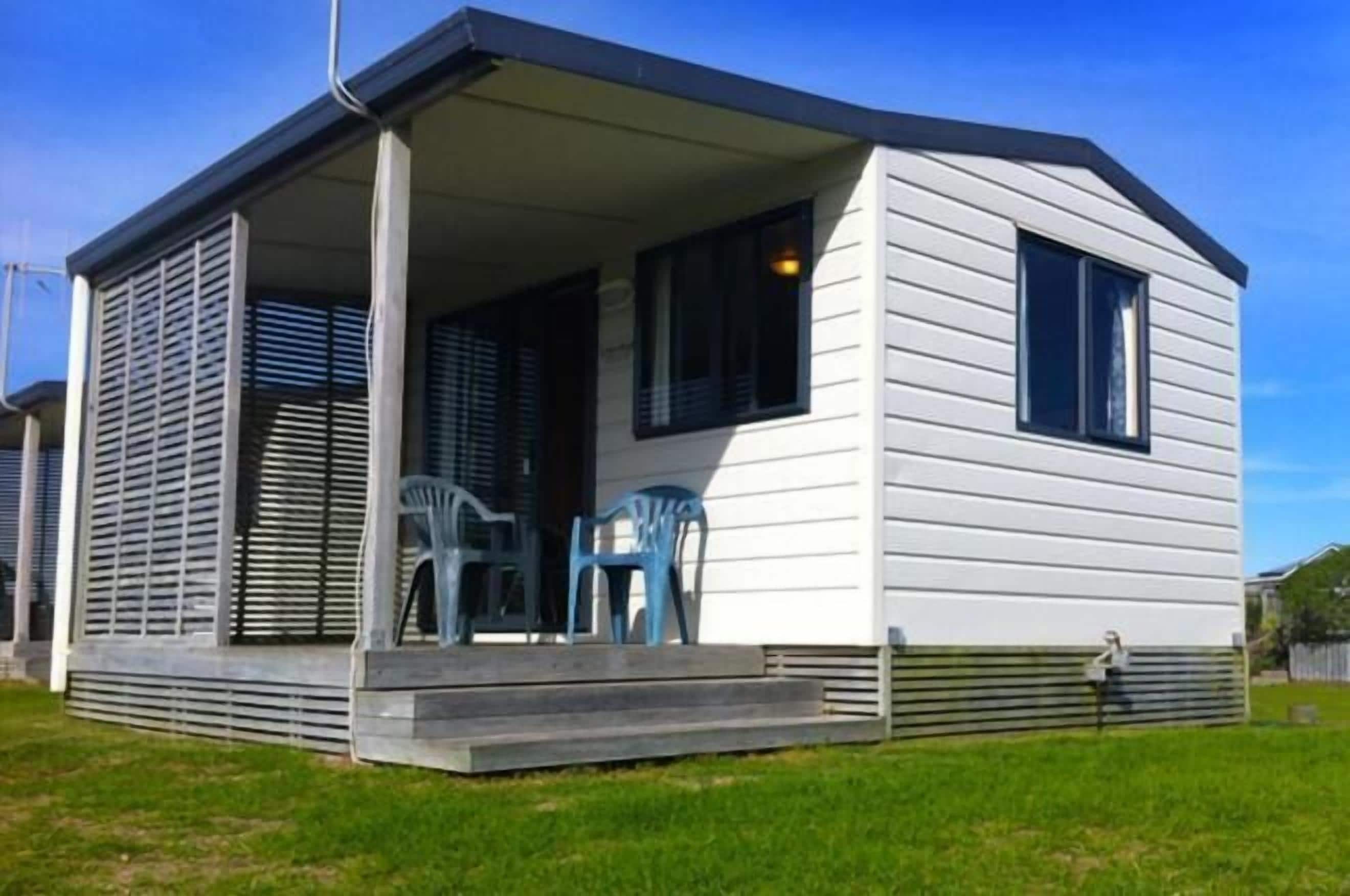 Una casa móvil de un piso con un porche, dos ventanas y una pequeña zona de estar al aire libre con sillas de plástico.