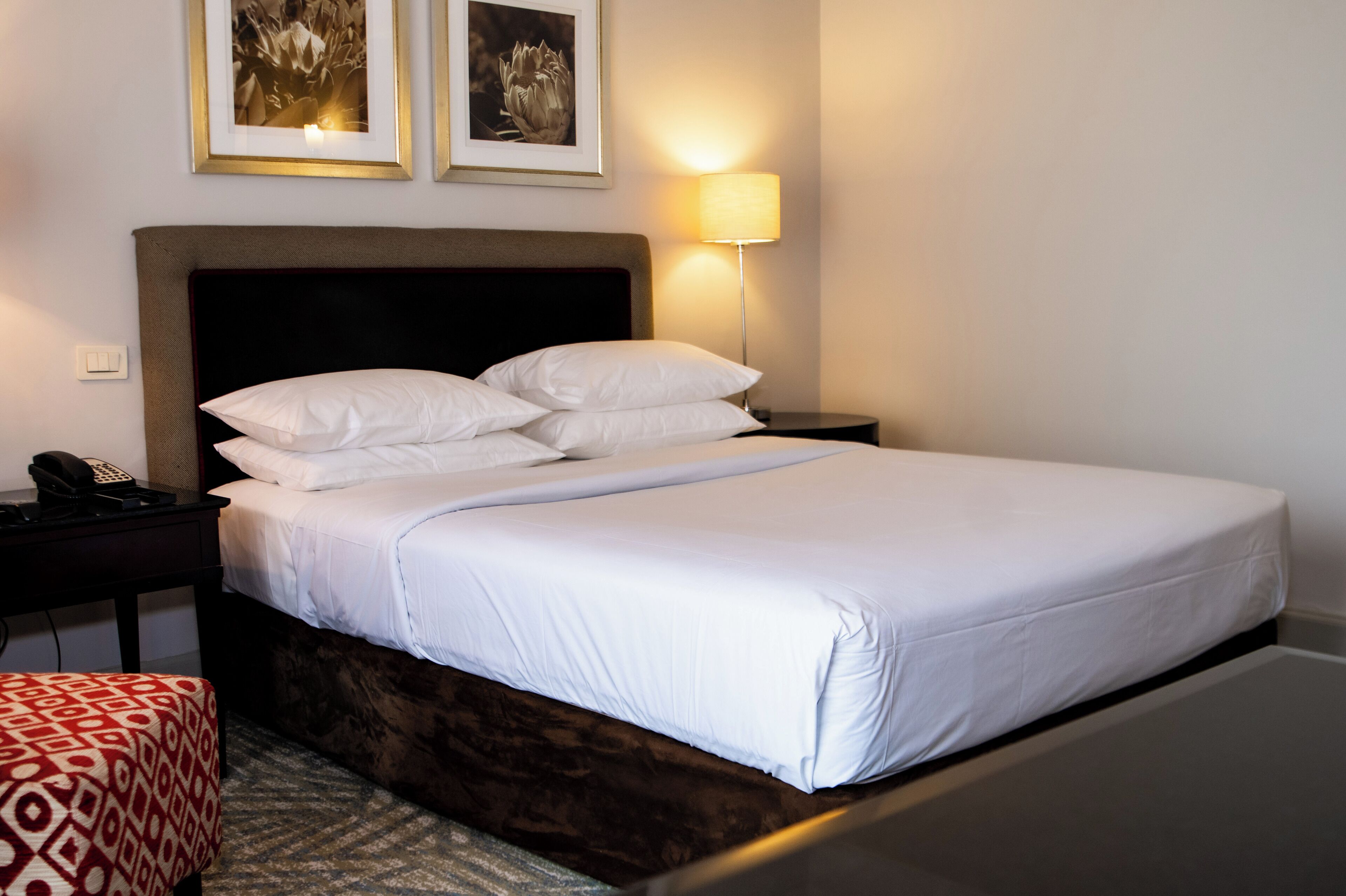 Een netjes opgemaakt bed met witte lakens, een donker hoofdbord en een bedlampje. Er staat een klein nachtkastje met een telefoon en een decoratief object.