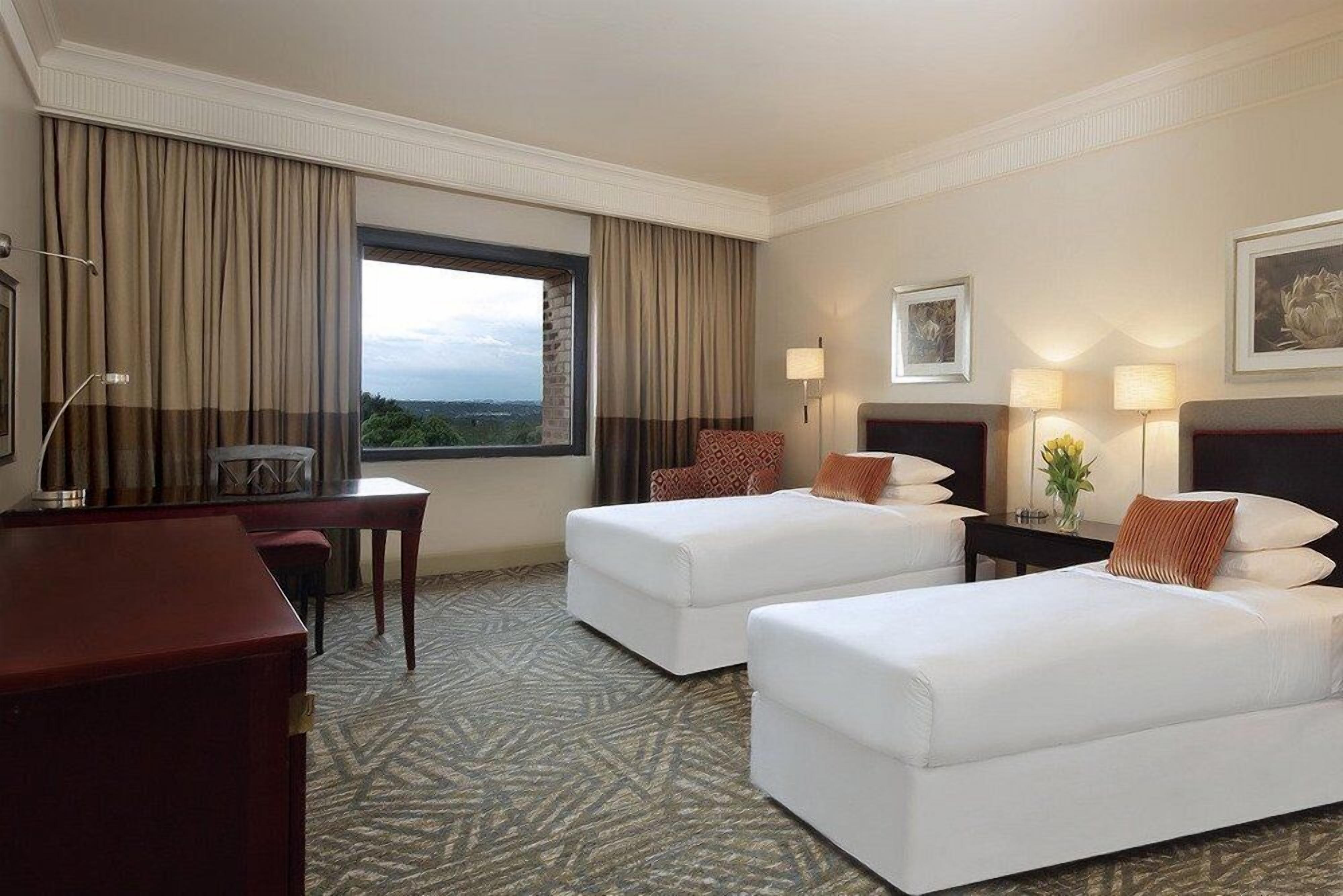 A hotel room with two beds, a desk, a chair, a lamp, and a view of the outdoors.