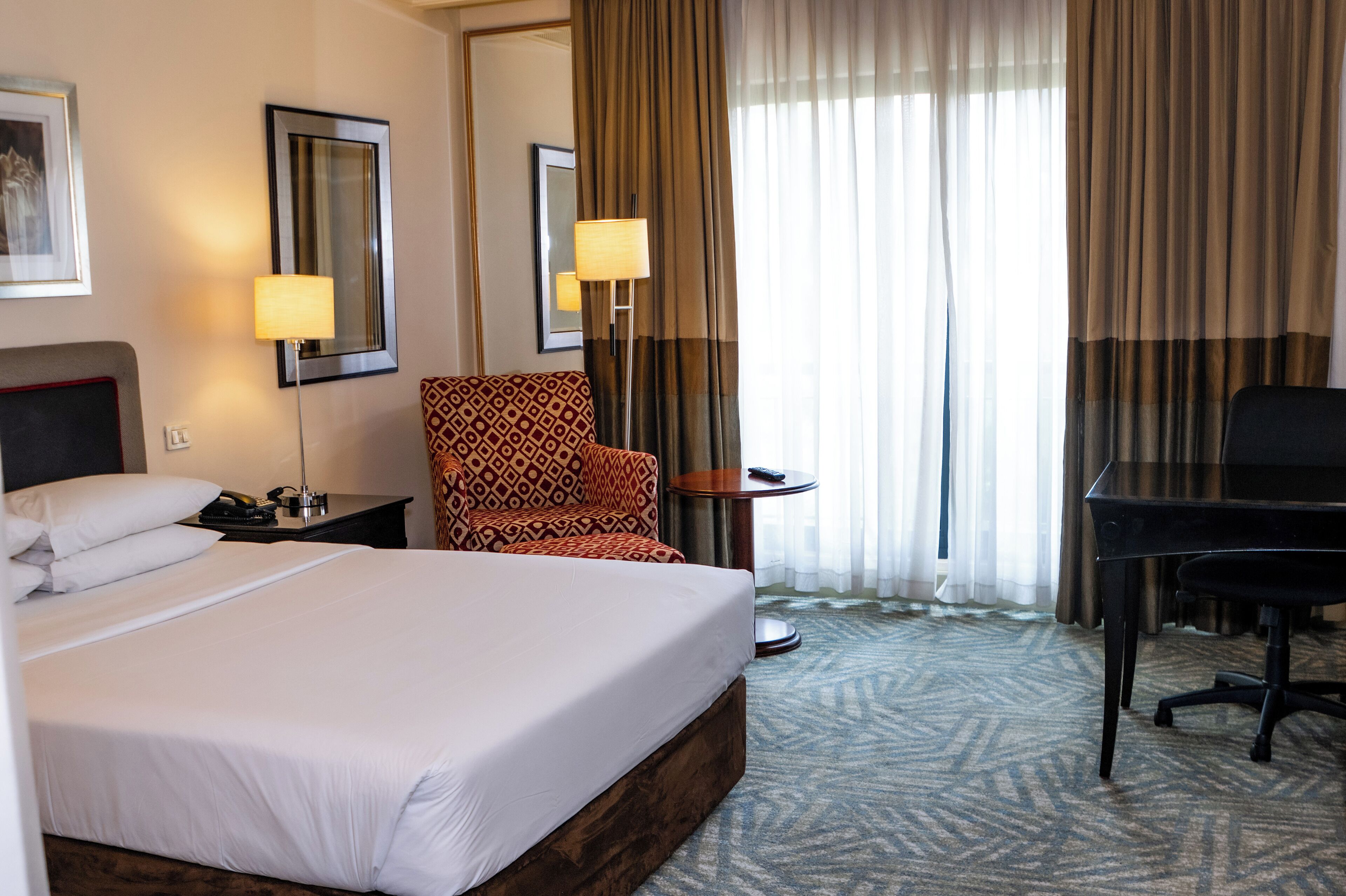 A hotel room with a large bed, a desk, a chair, a lamp, and a mirror.