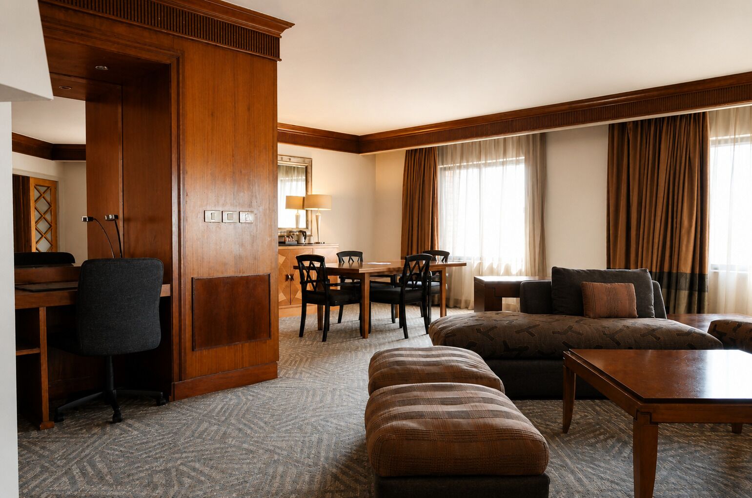 A hotel room with a wooden wardrobe, a dining area with a table and chairs, a sofa, an ottoman, and a small wooden table.
