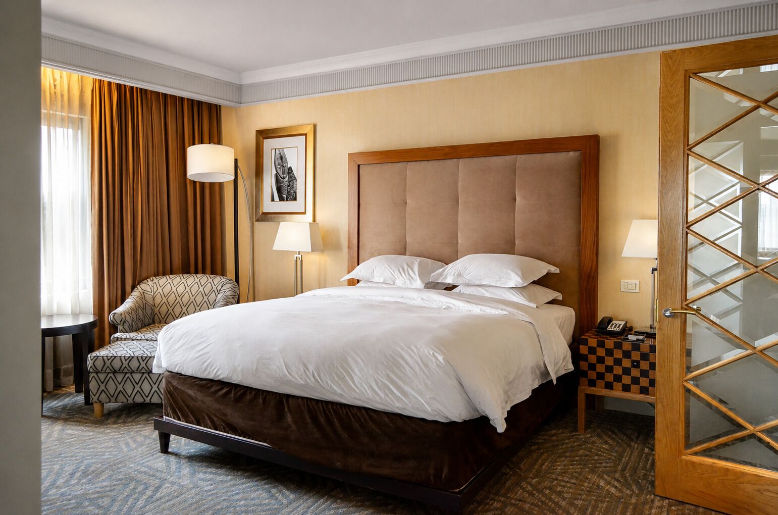 A hotel room with a large bed, a chair, a desk, and a door with glass panels.