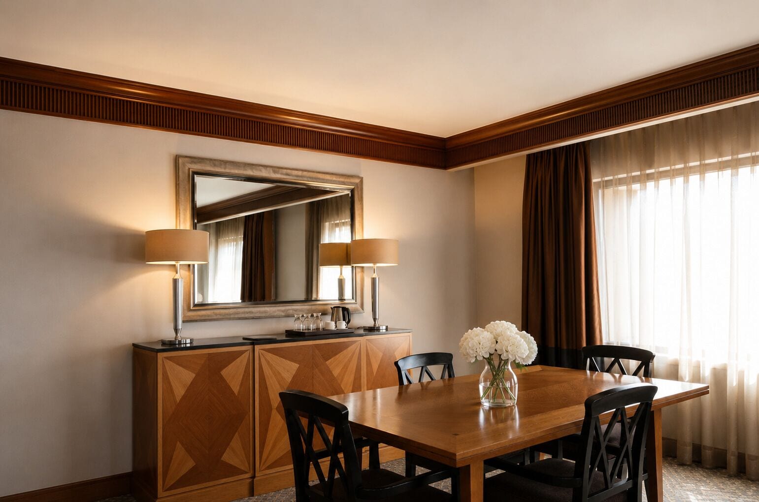 A hotel dining room with a wooden table, four chairs, a large mirror, two lamps, and a vase with flowers.