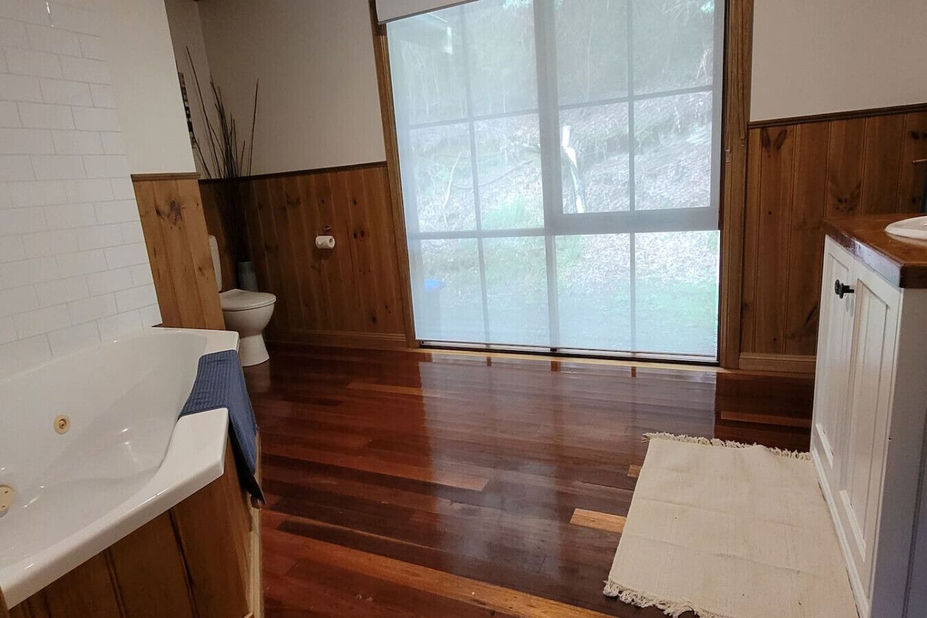 Une salle de bain avec un plancher en bois, un lavabo blanc, des toilettes et une grande fenêtre.