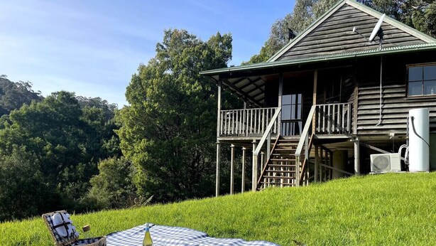 A wooden house with a balcony, a picnic setup on a grassy hill, and a clear sky. - Tall Trees Eco Retreat - Manna Gum Cottage (Apollo Bay)