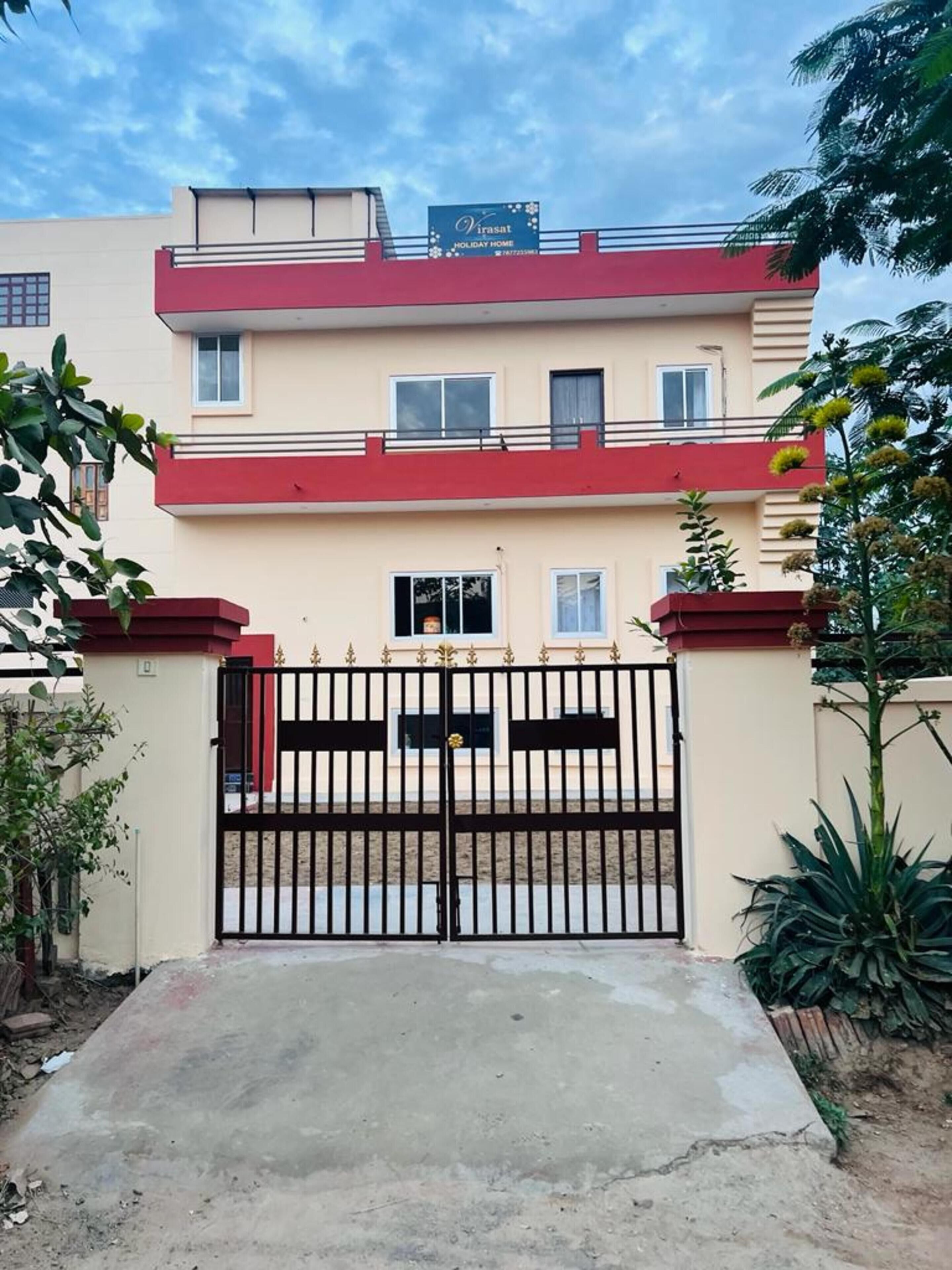 A two-story building with a red and beige facade, a gated entrance, and a sign indicating 'Virasat Holiday Home'.