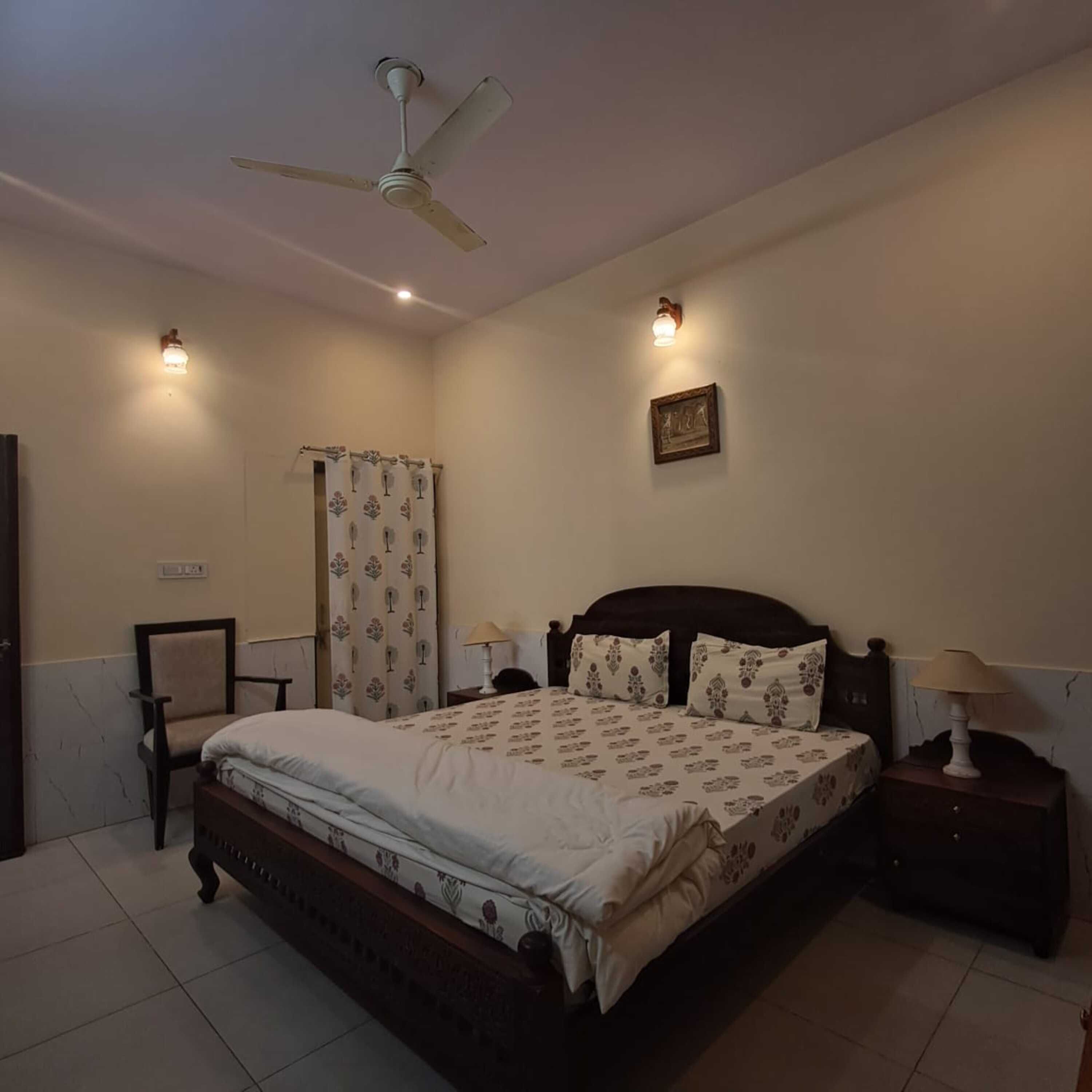 A bedroom with a bed, a nightstand, a chair, a ceiling fan, and a wall-mounted light fixture.