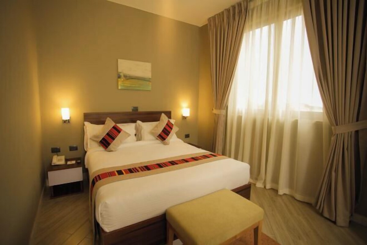 Habitación de hotel con una cama grande, lámparas de noche, una mesita de noche, una ventana con cortinas y un cuadro en la pared.