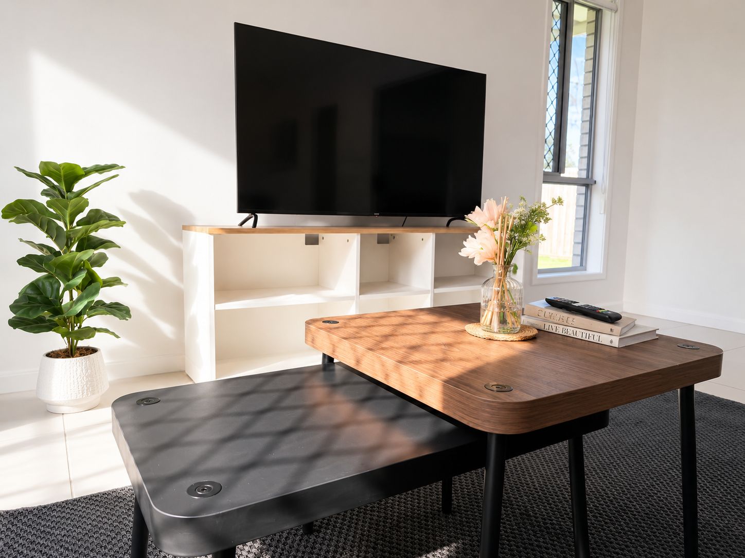 Ein modernes Wohnzimmer mit einem Flachbildfernseher, der an einem weißen Regal befestigt ist, einem Holztisch mit einer Bank, einer Topfpflanze und einer Vase mit Blumen.