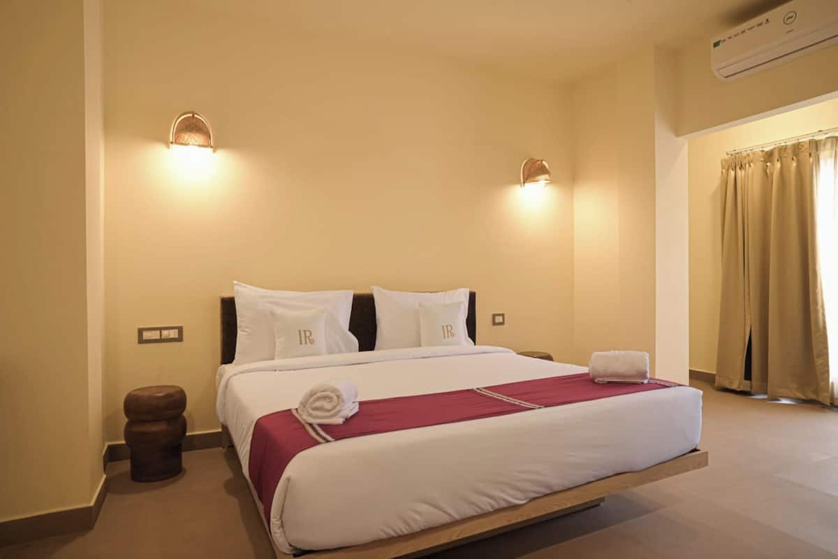Une chambre d’hôtel avec un grand lit, deux lampes fixées au mur, une tête de lit en bois, une petite table de chevet, une poubelle marron et des rideaux beiges.