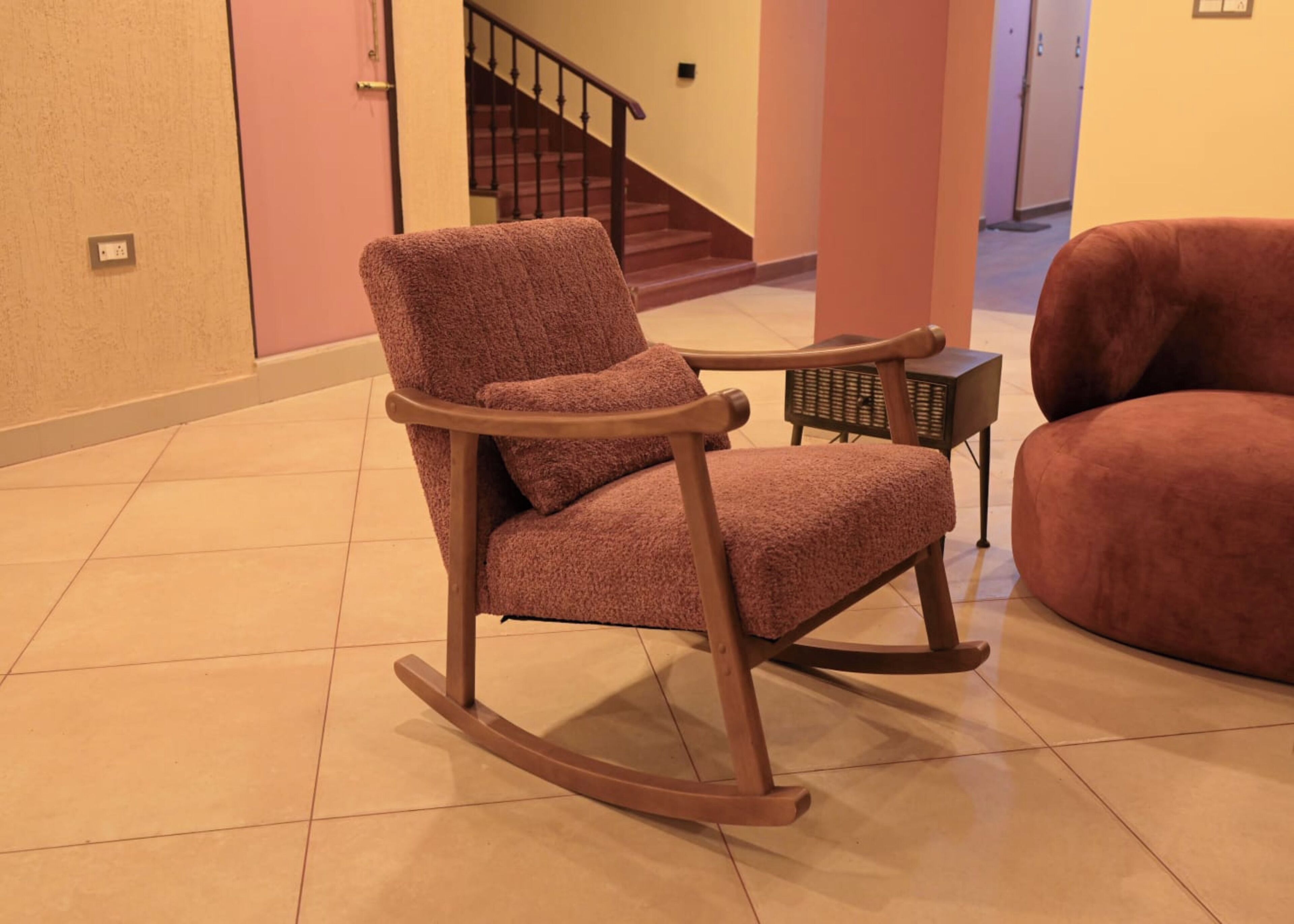 A rocking chair in a room with a tiled floor, a staircase in the background, and a small table with a basket.