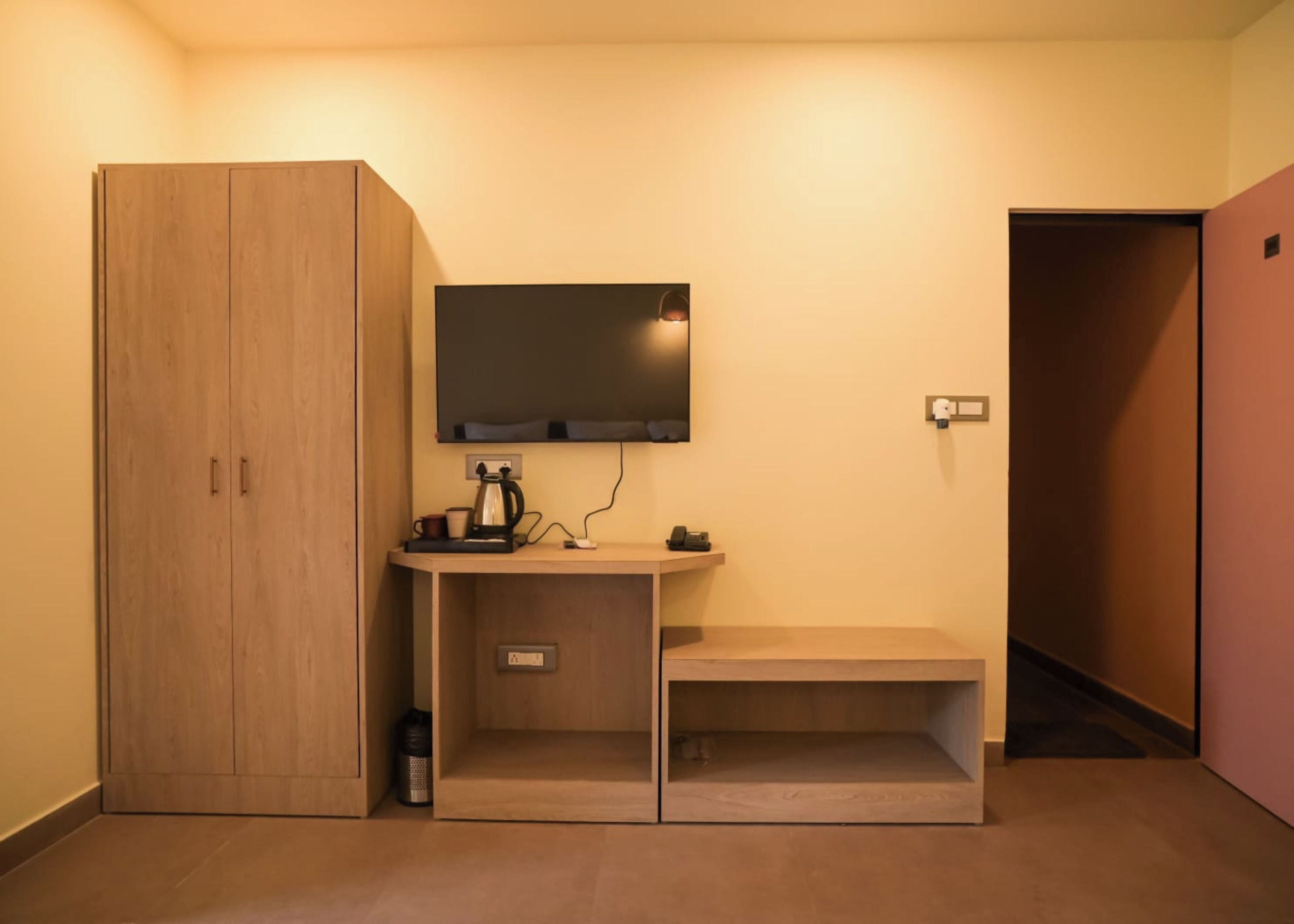 A small room with a wooden wardrobe, a desk with a kettle, a phone, and a wall-mounted TV.