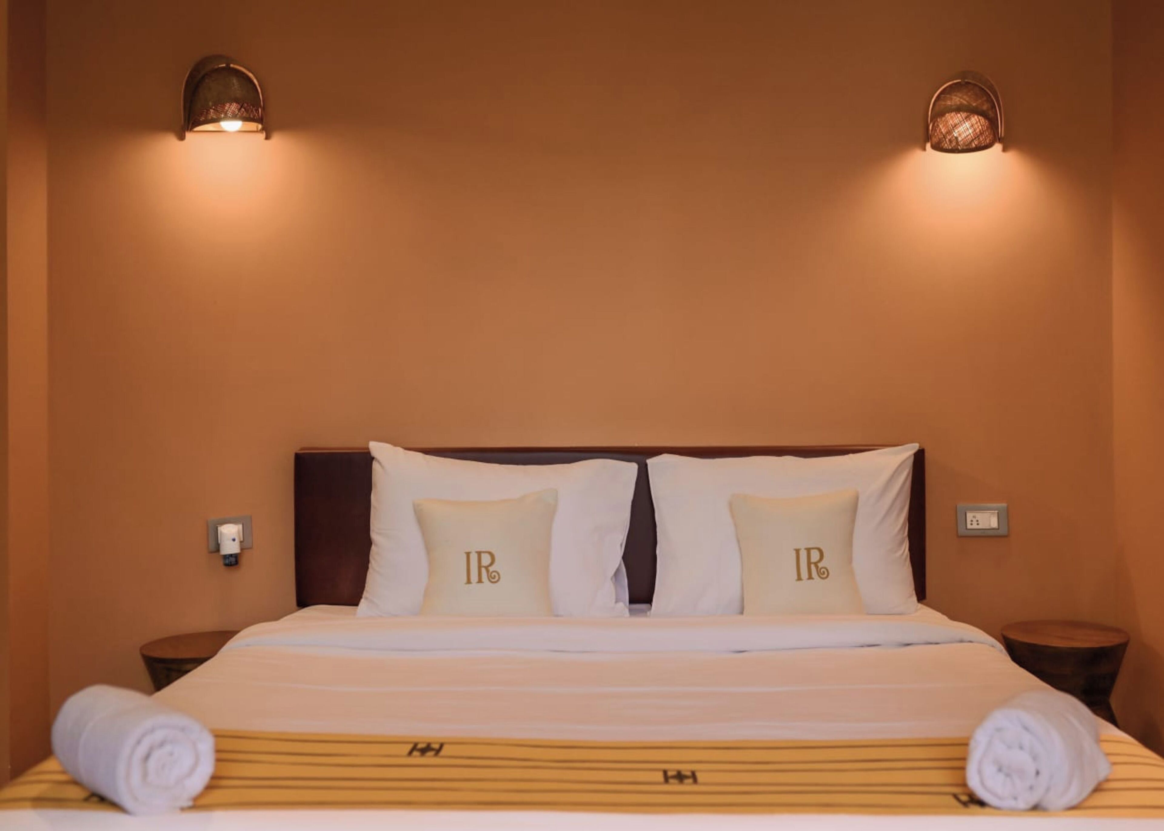 Habitación de hotel con cama, dos almohadas con las iniciales 'IR', cabecera, dos luces fijadas a la pared y mesita de noche con una toalla.