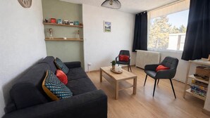Una sala de estar con sofá, dos sillones, una mesa de centro, una estantería con adornos y una ventana con cortinas.