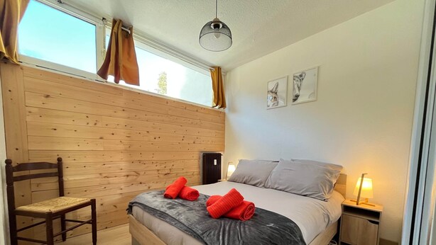 Un dormitorio con piso de madera, una cama con ropa de cama gris y beige, dos toallas rojas sobre la cama, una silla de madera, una mesita de noche con lámpara y una ventana con cortinas.