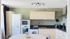 Una cocina compacta con microondas, horno y tostadora. Hay una mesa de comedor con sillas y un frutero. La cocina tiene armarios de madera clara y una encimera negra.