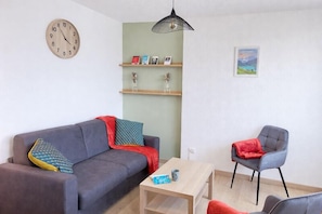 Un salon comprenant un canapé, un fauteuil, une table basse, une horloge, des étagères ornées d’objets décoratifs et un luminaire suspendu.