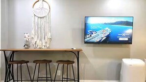 A bar area with a wooden table, three stools, a macrame wall hanging, and a television displaying a cruise ship. - Private Urban Oasis Near SF - w/kitchenette 22421B (Hayward)