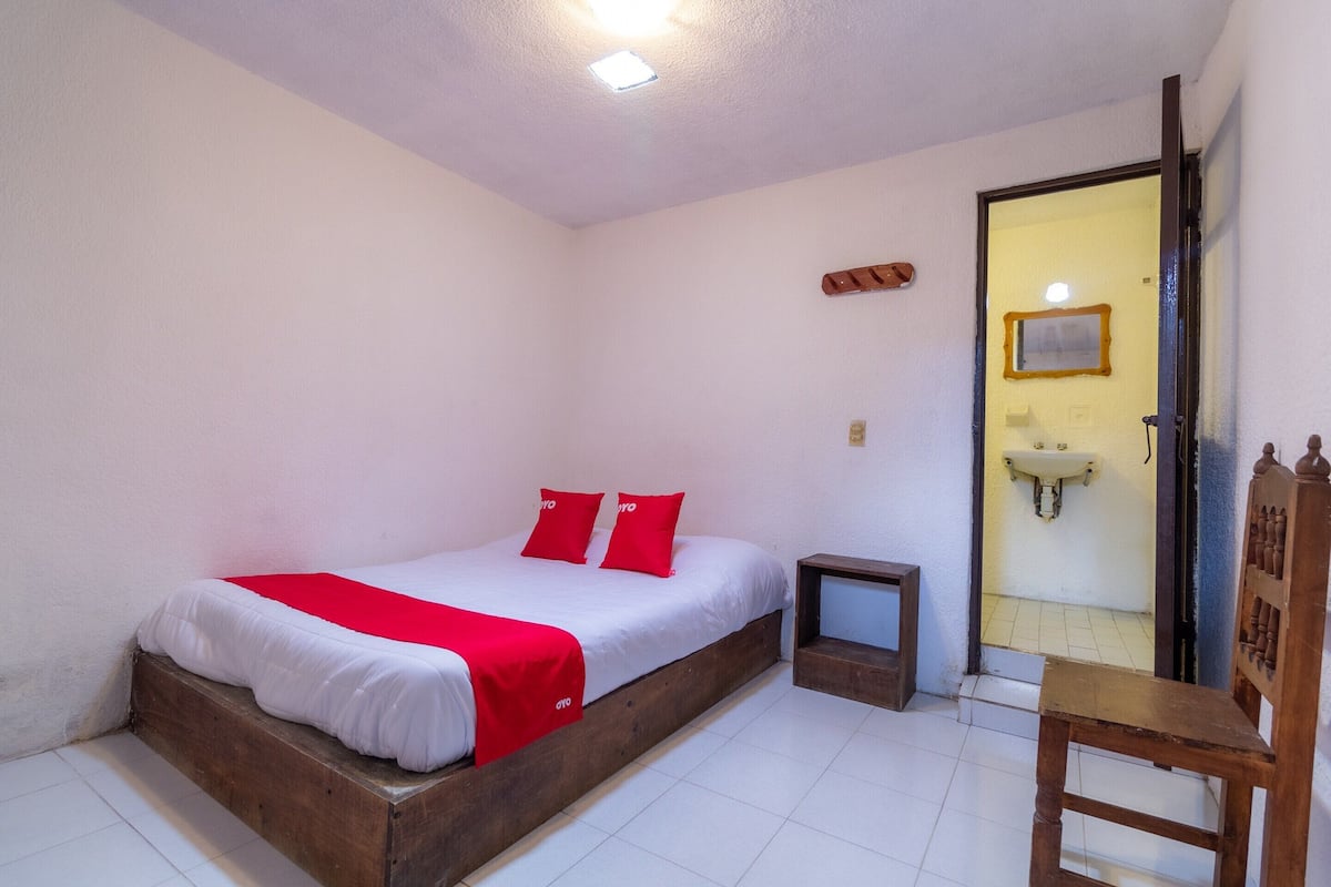 A single bed with a wooden frame, a bedside table, a chair, a small bedside table, and a door leading to a bathroom.