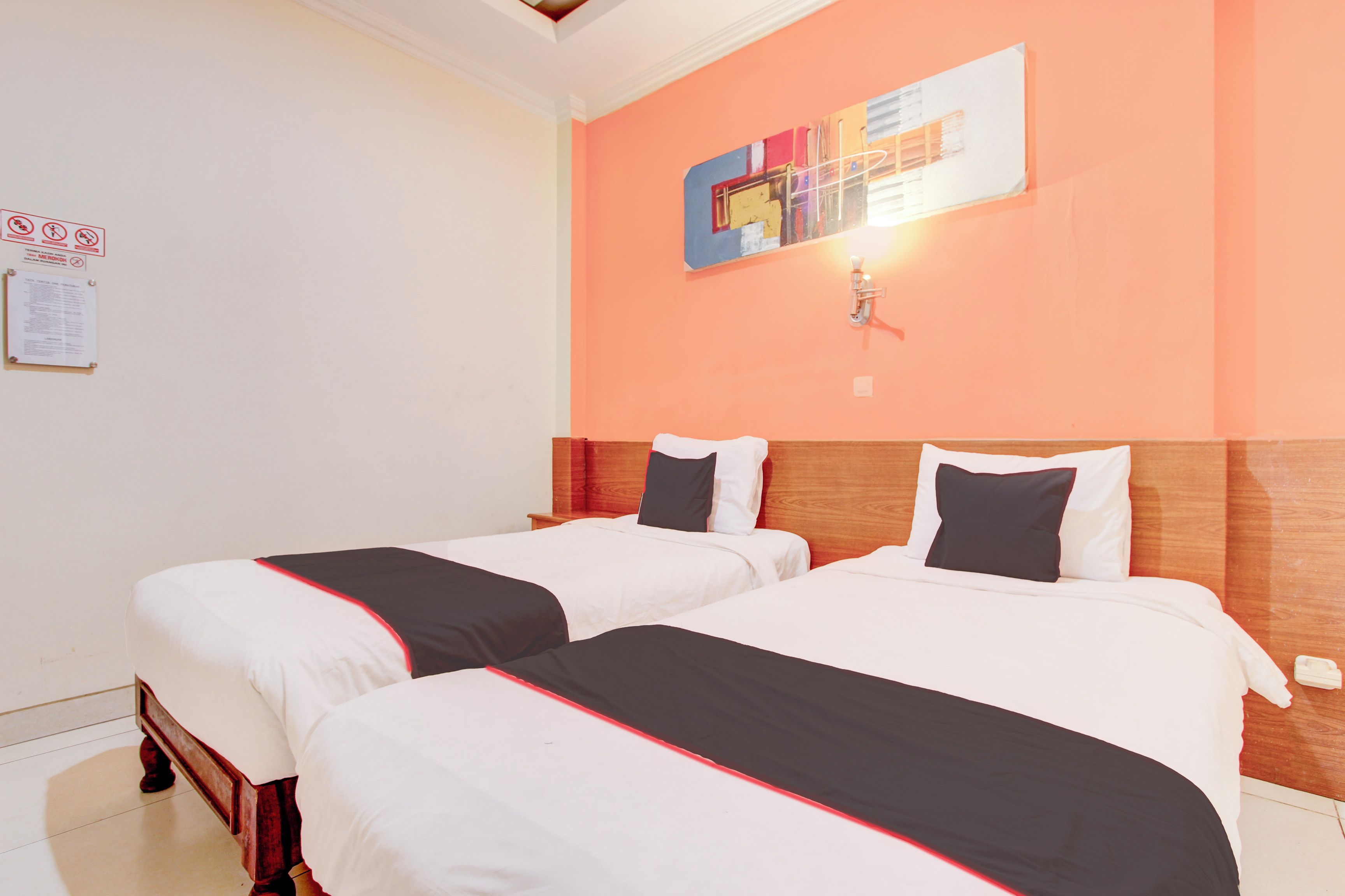 Habitación de hotel con dos camas, ropa de cama blanca con detalles rojos, cabecera de madera, una obra de arte colgada en la pared y un interruptor de luz.