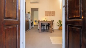 Une porte en bois donne sur un coin repas, où se trouvent une table et des chaises. Un climatiseur est fixé au mur, et une plante en pot est posée à proximité.