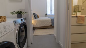 Un espace buanderie avec un lave-linge et un sèche-linge, un lit près d’une fenêtre et une petite plante sur la commode.