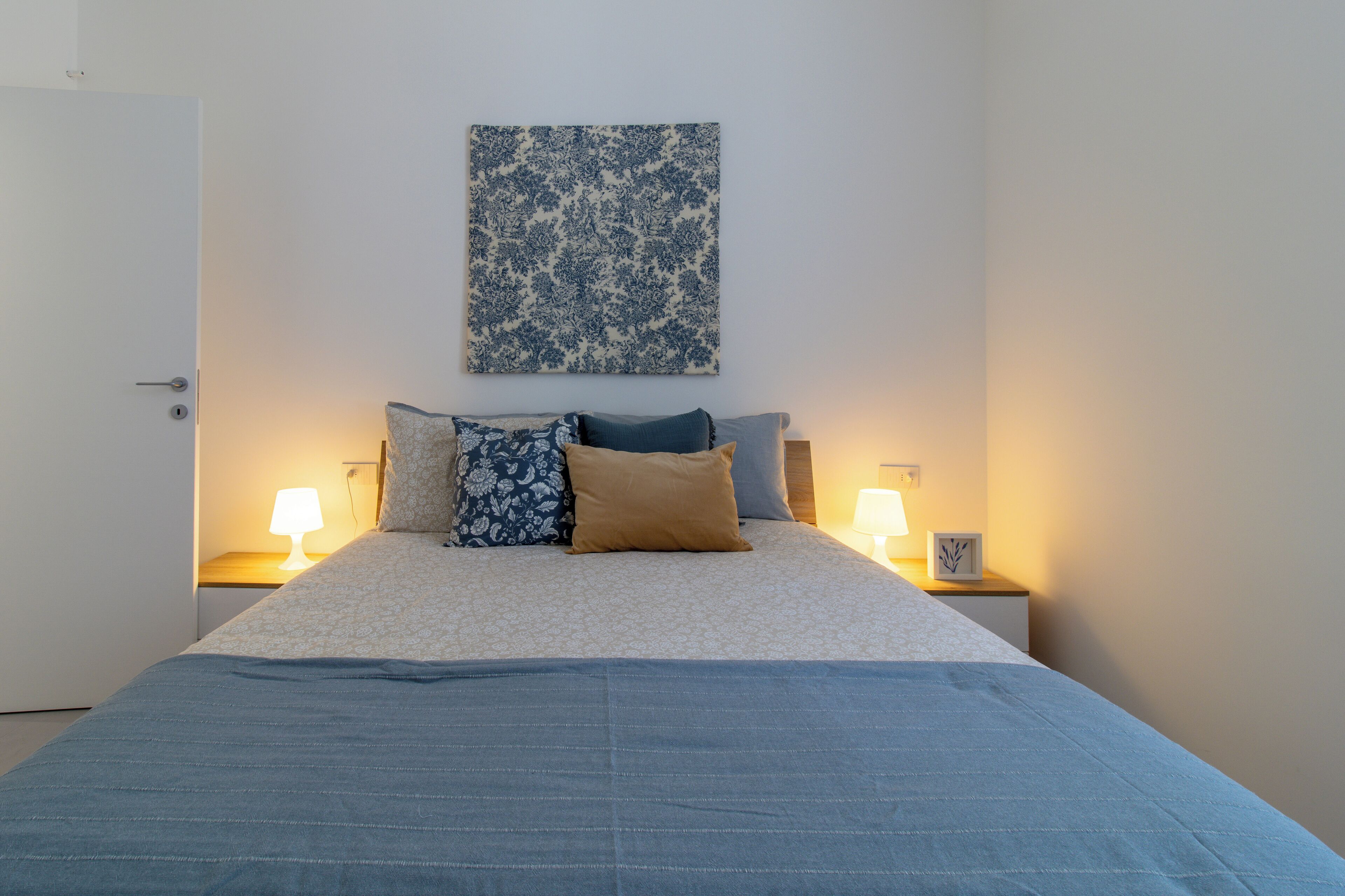 A neatly made bed with a blue bedspread, two bedside lamps, and a floral-patterned wall hanging.