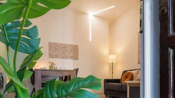 Un salon moderne avec un canapé, une table basse et une lampe. On y trouve une porte sur laquelle est posé un paillasson avec l’inscription « Bienvenue ».