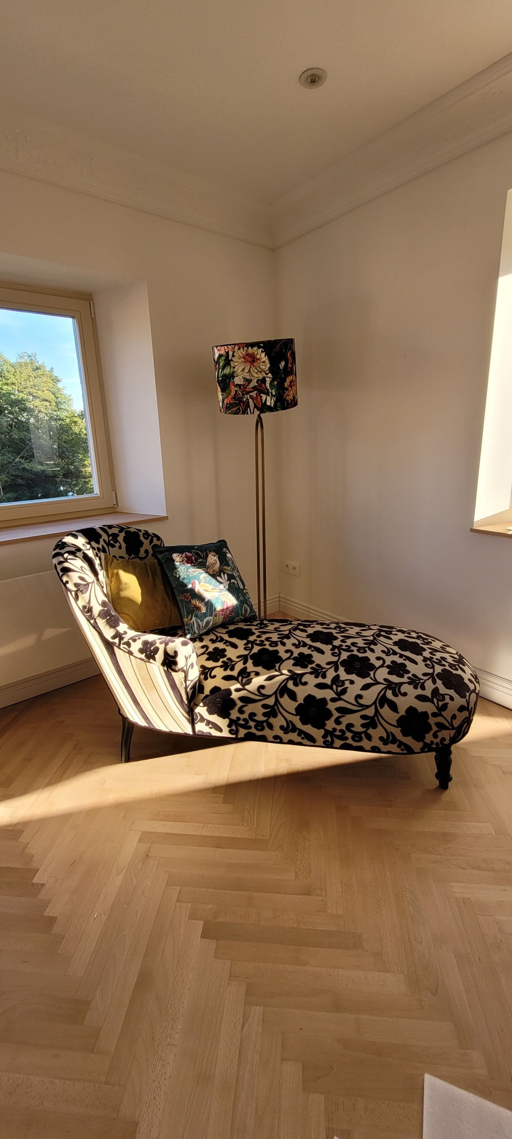 Ein Zimmer mit einer gepolsterten Couch, einer Stehlampe und einem Fenster mit Blick auf Bäume.