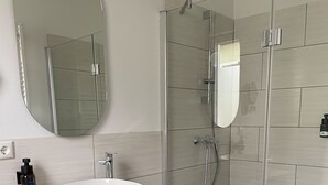 A modern bathroom with a large oval mirror, a white sink on a wooden vanity, black drawers, and a glass shower enclosure. - Das Hartmanns (Pegnitz)