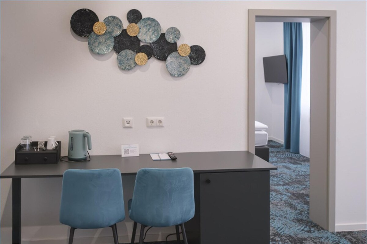 A hotel room with a dark desk, two blue chairs, a decorative wall piece, a kettle, and a small table with a tray.