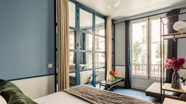 A hotel room with a bed, a small table with a vase of flowers, a chair, and a window with curtains. - Merveil Luxury Suite - Saint Denis I (Paris)