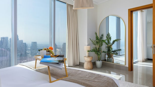 A modern bedroom with a large bed, a bedside table with magazines, a potted plant, and a view of the city through large windows.