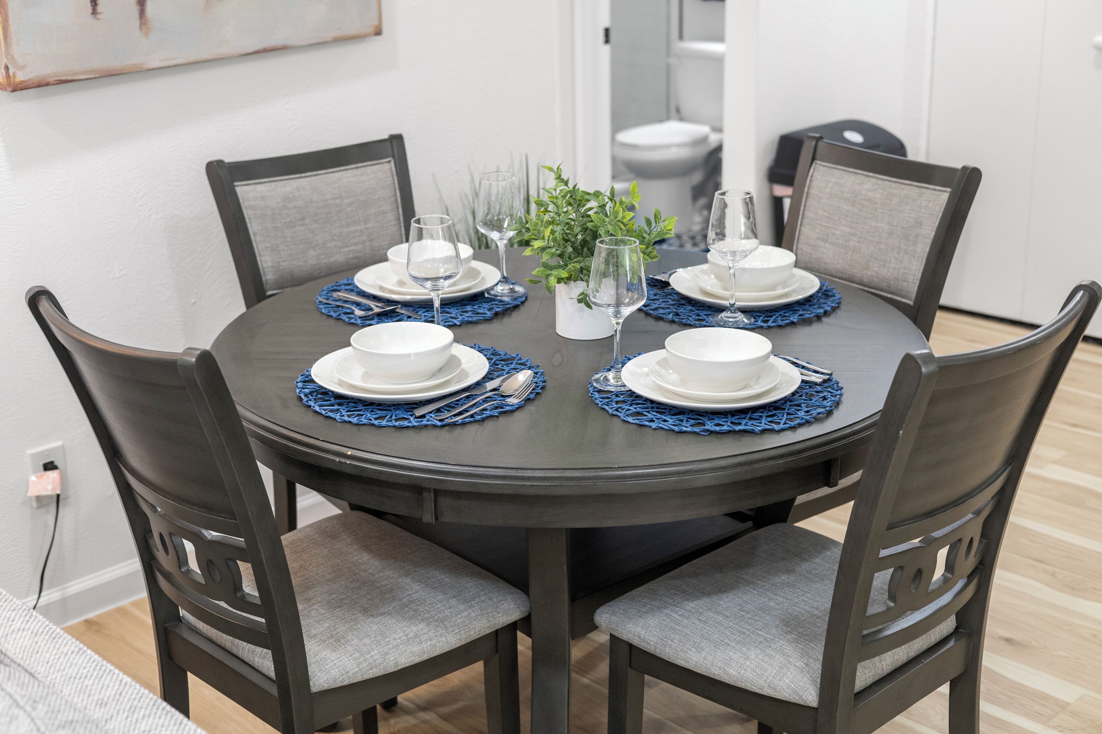 A round dining table with six chairs, each with a cushion. The table is set with white plates, silverware, and blue placemats. A small potted plant is in the center.
