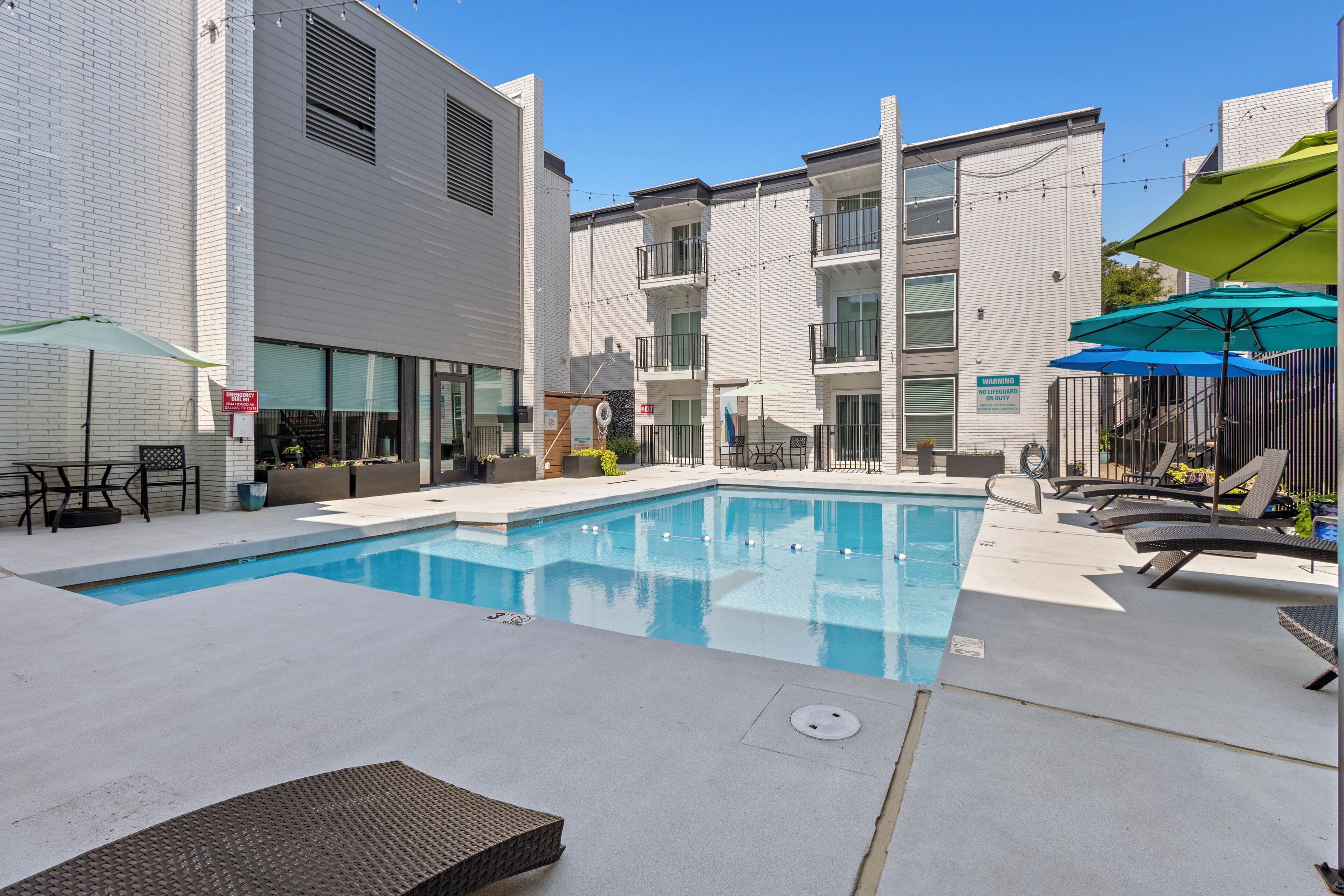 A swimming pool with lounge chairs and umbrellas, surrounded by white buildings with balconies.