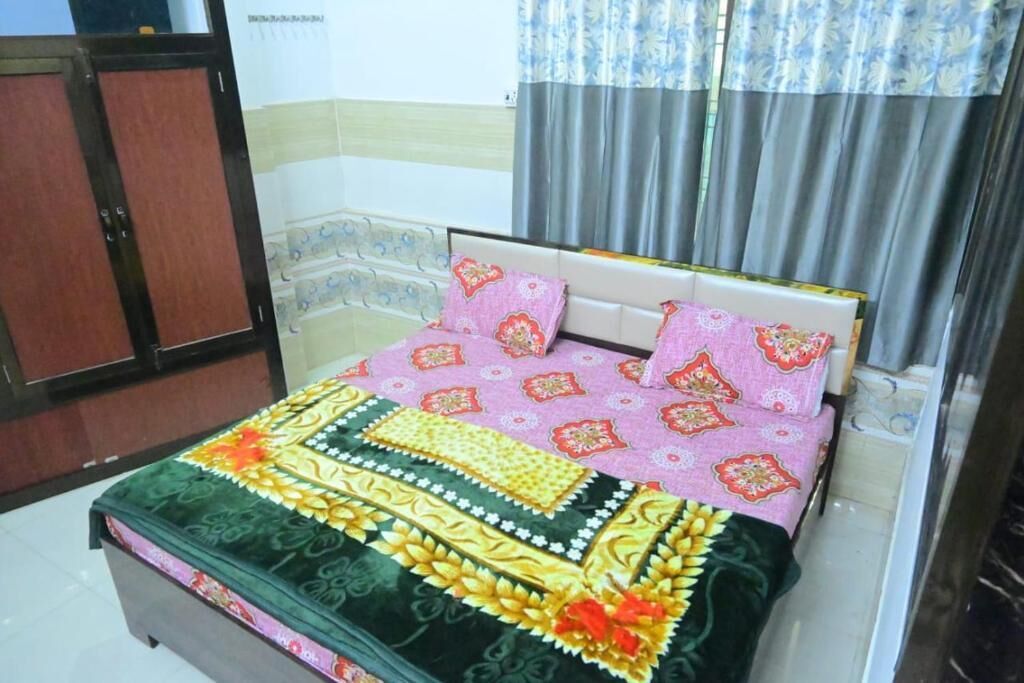 A neatly made bed with a patterned bedspread and pillows, a wooden wardrobe, and a window with curtains.