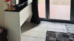 Une pièce avec un téléviseur posé sur un meuble blanc, un miroir au-dessus, et une vue sur un jardin par une porte coulissante.