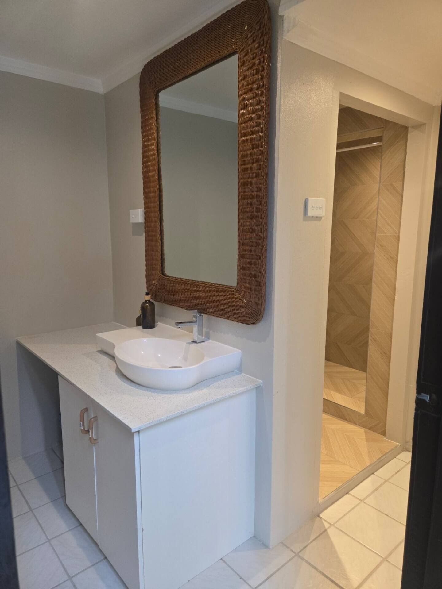 A bathroom with a white sink, a wicker-framed mirror, and a shower area with wooden flooring.